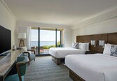 A hotel room with two beds, a TV, a desk, and a view of the sea.