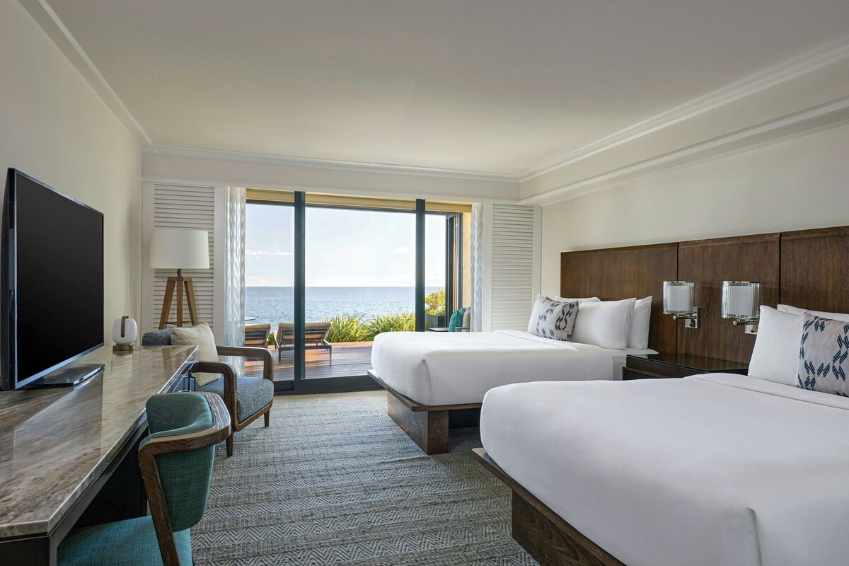 A hotel room with two beds, a TV, a desk, and a view of the sea.