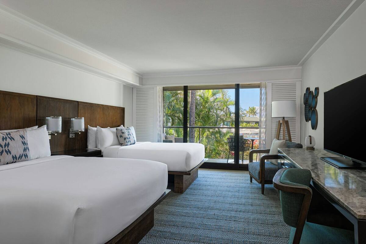 Une chambre d’hôtel avec deux lits, un bureau, une télévision et une vue sur les palmiers.