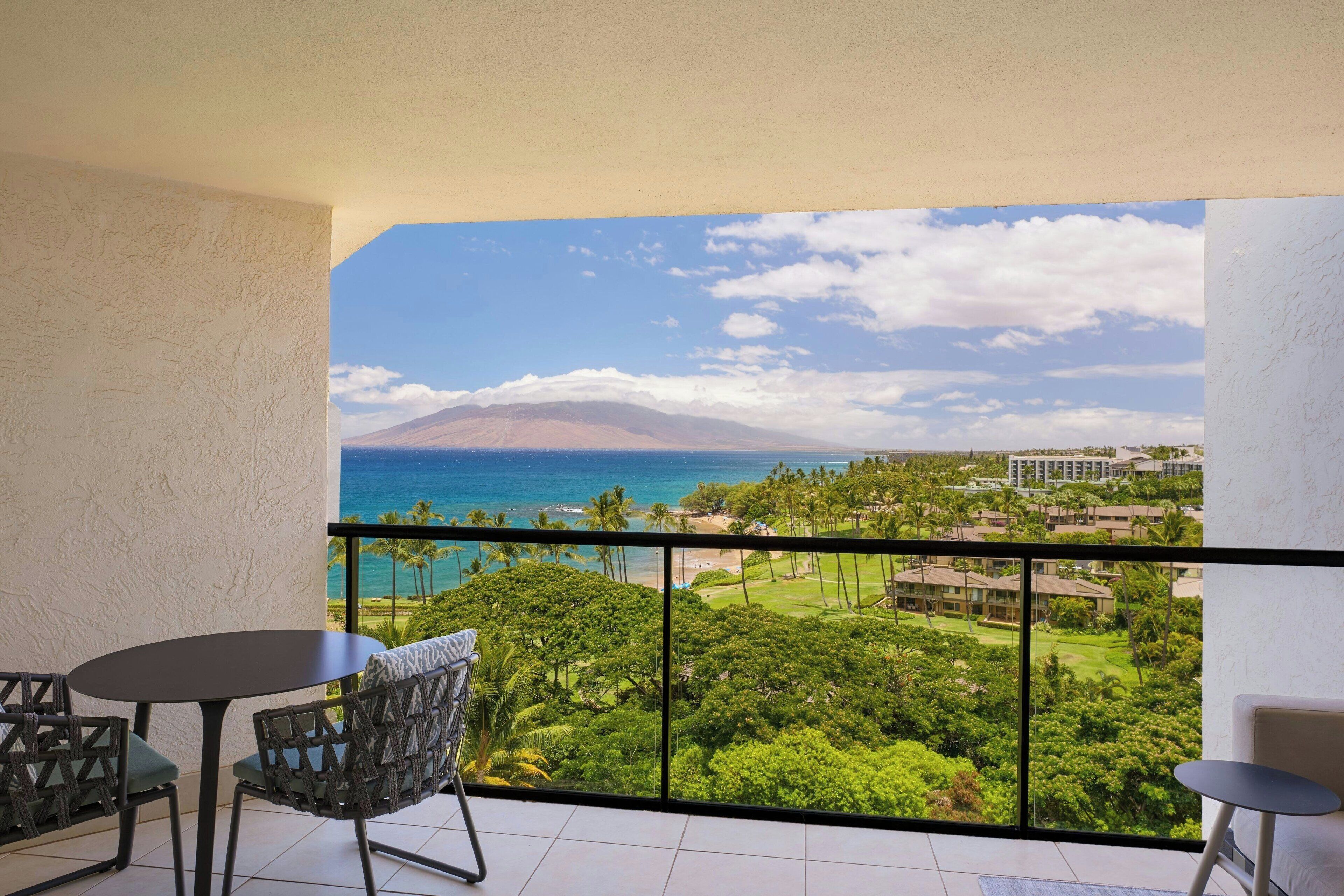 A balcony with a view of the ocean and a mountain, furnished with two chairs and a small table.