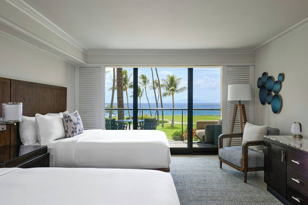 A hotel room with two beds, a desk, a chair, and a view of the ocean.