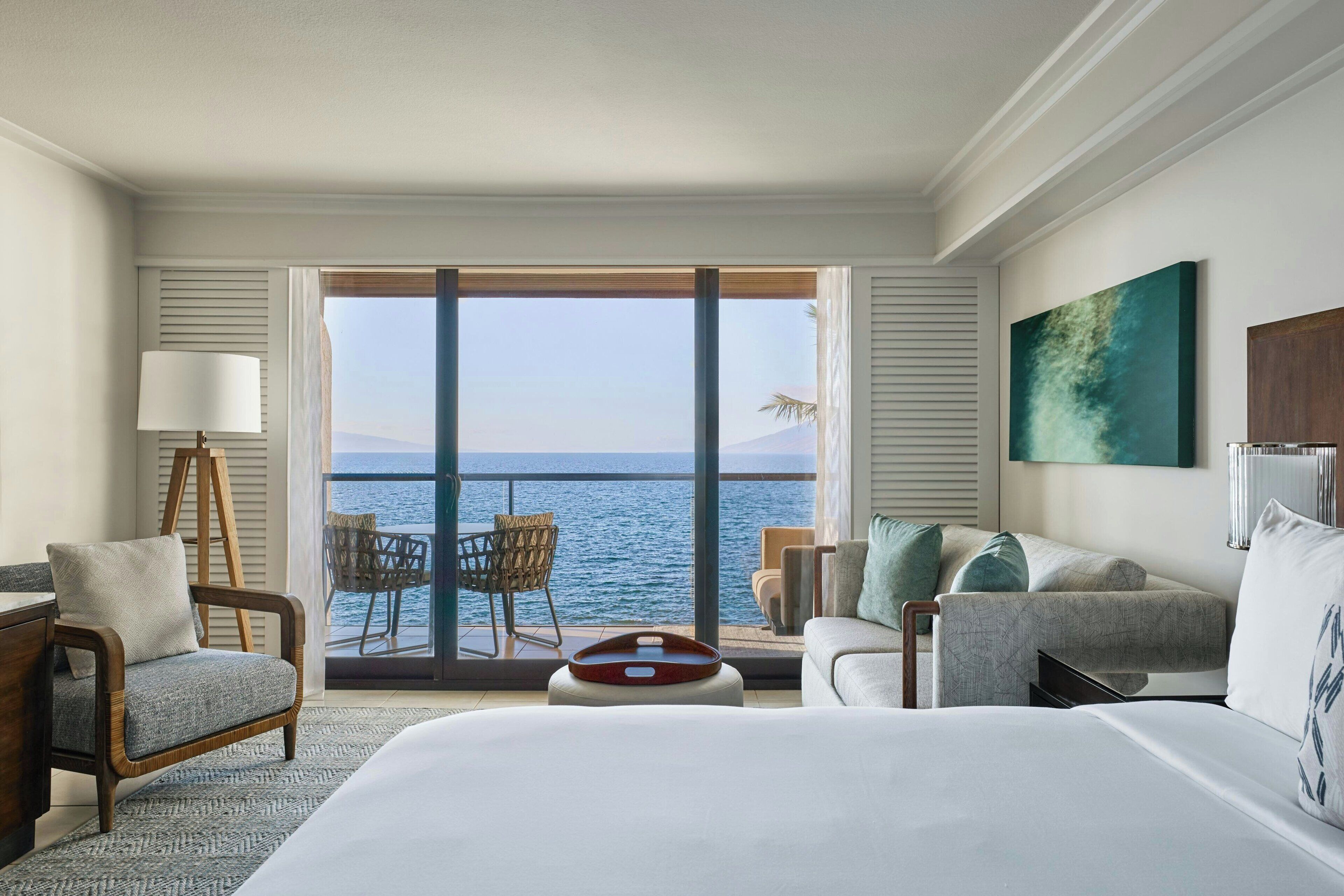 A hotel room with a large bed, a desk, a chair, a small table, and a view of the sea through a sliding door.