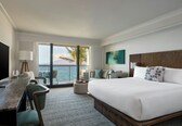 A hotel room with a large bed, a desk with a chair, a view of the ocean, and a balcony with chairs.