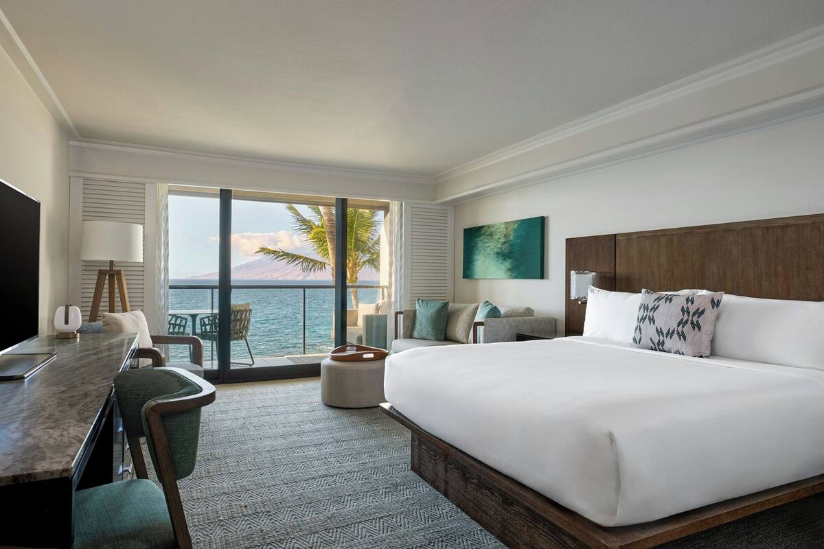 A hotel room with a large bed, a desk with a chair, a view of the ocean, and a balcony with chairs.
