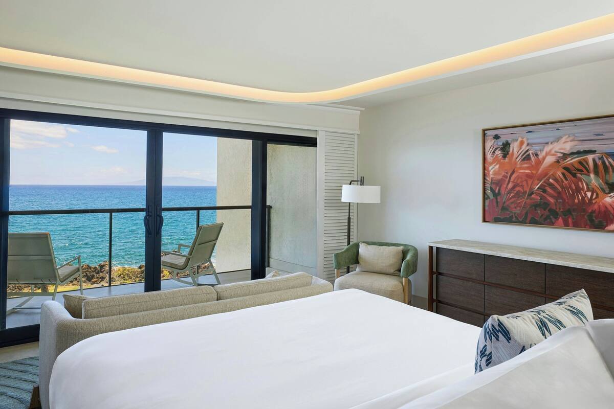 A hotel room with a large bed, a seating area with chairs, a painting, and a view of the ocean.