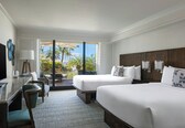 A hotel room with two beds, a desk, and a TV. There is a view of palm trees and a pool outside.