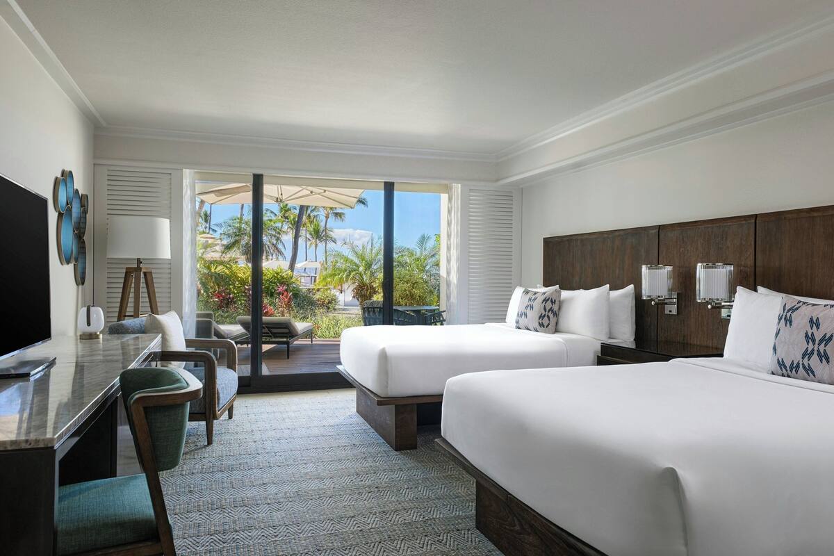 Une chambre d’hôtel avec deux lits, un bureau et une télévision. La fenêtre offre une vue sur des palmiers et une piscine.