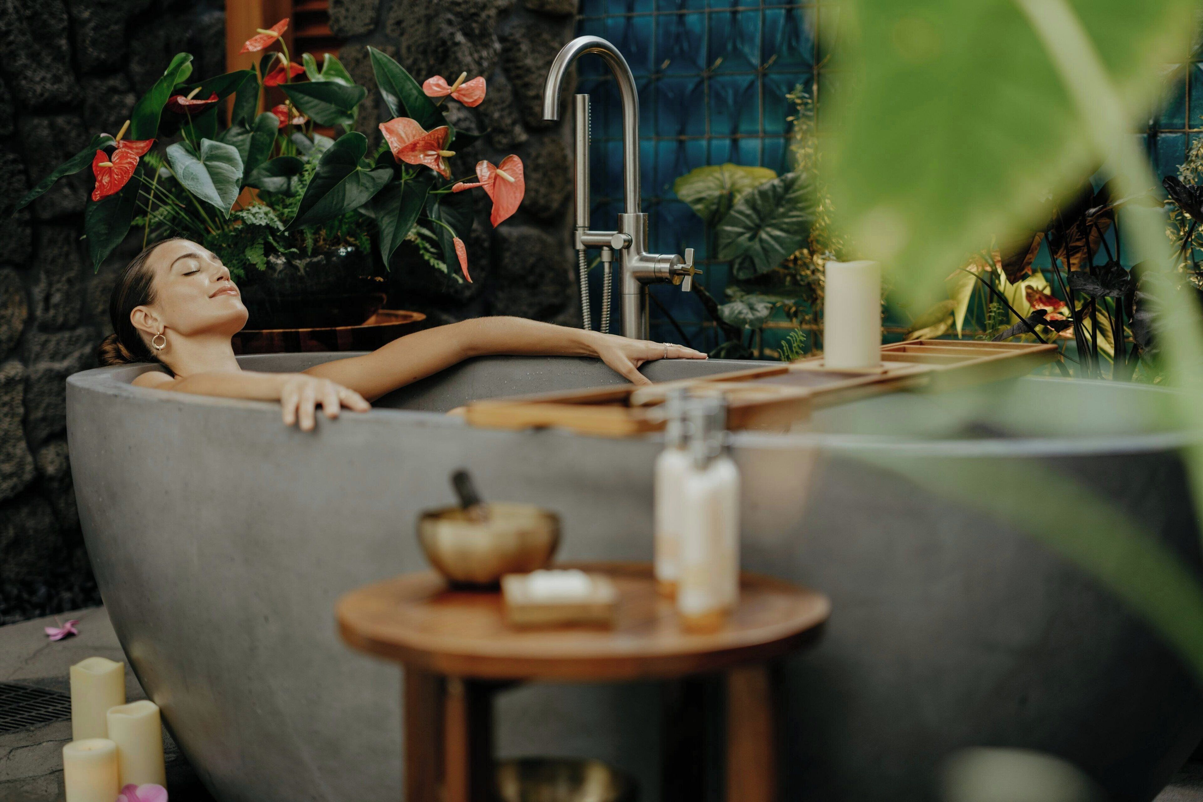 A person relaxing in a bathtub with a modern faucet, surrounded by lush plants and candles.