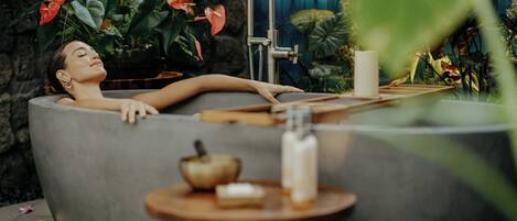 Une personne se détend dans une baignoire, avec un robinet moderne, entourée d’une végétation luxuriante et de bougies.