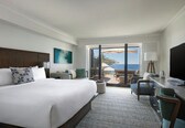 A hotel room with a large bed, a desk, a TV, and a balcony with a view of the ocean.