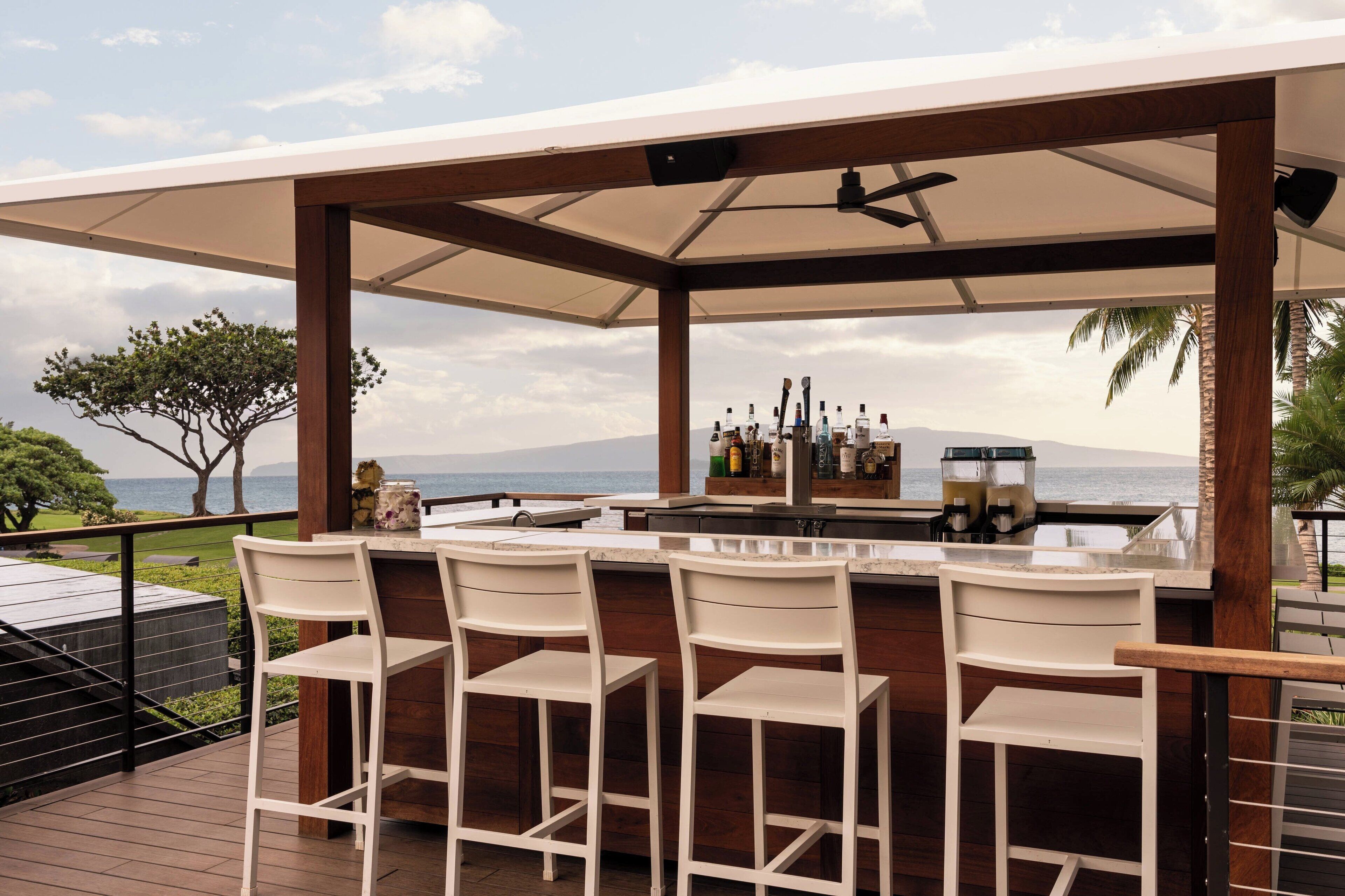 A covered outdoor bar area with a wooden deck, white bar stools, and a view of the sea.