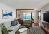 A modern living room with a sofa, armchair, and coffee table, a TV, and a view of the ocean.