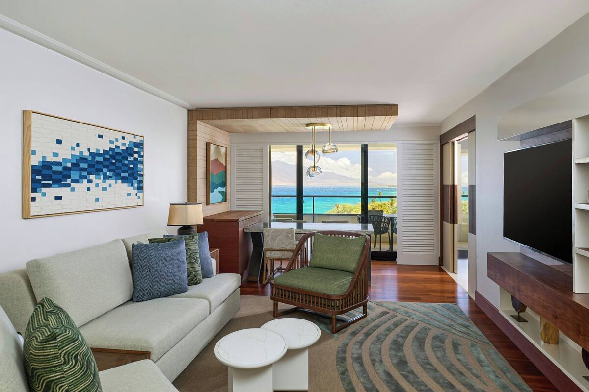 A modern living room with a sofa, armchair, and coffee table, a TV, and a view of the ocean.