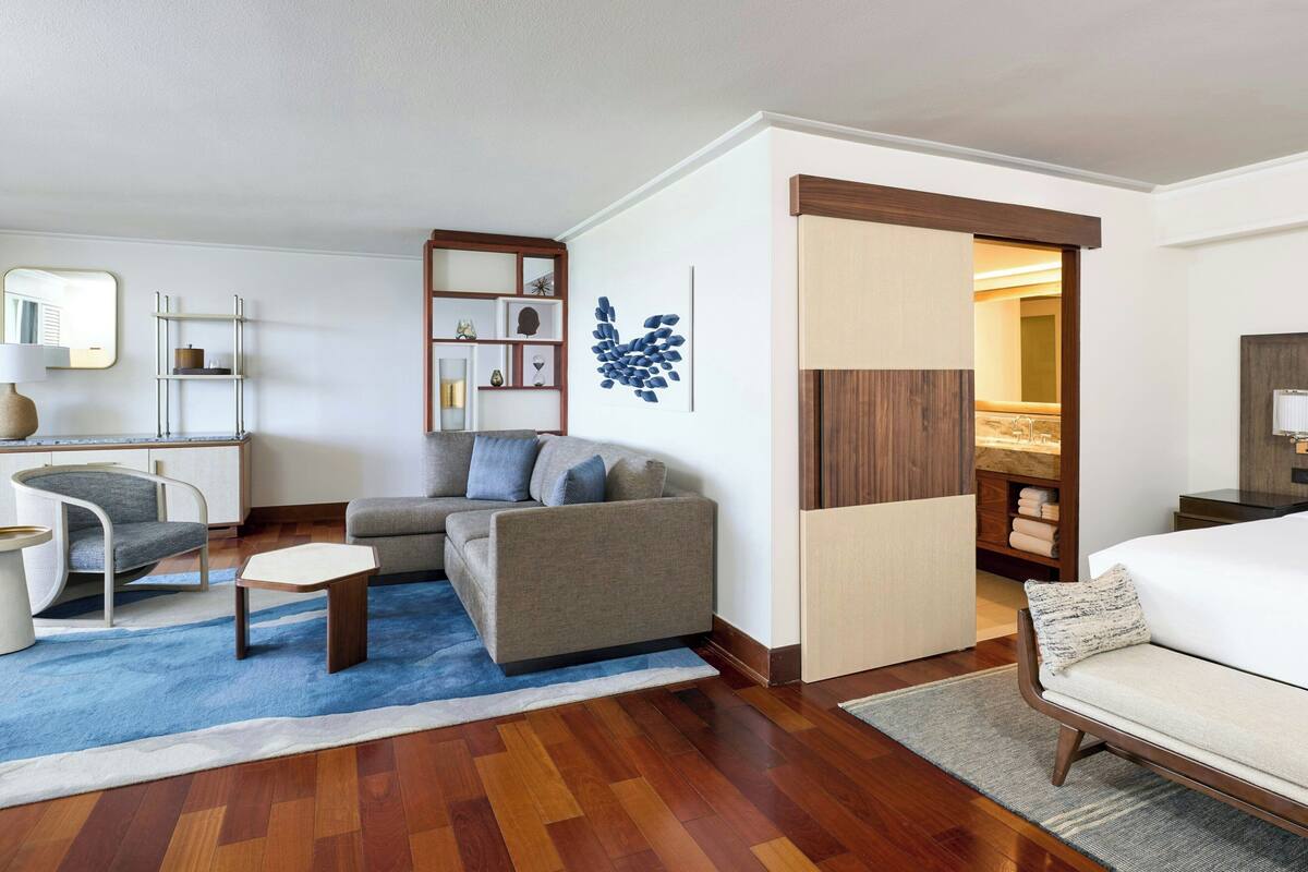 Une chambre d’hôtel moderne avec un canapé, un fauteuil et une petite table basse. Le sol est en bois et il y a un tapis bleu.