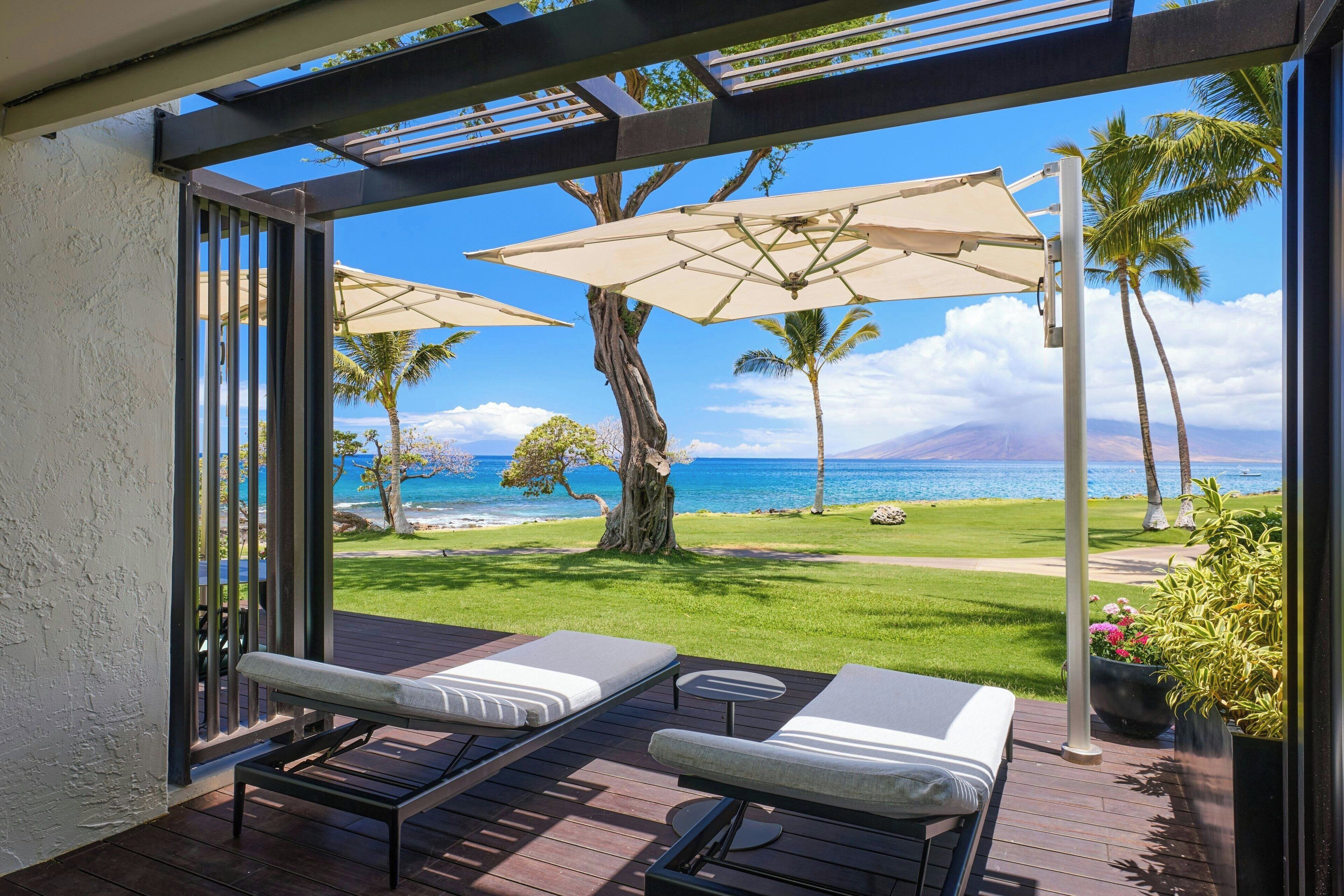 Eine Terrasse mit zwei Liegestühlen und einem Tisch, mit Blick auf einen Strand mit Palmen und den Ozean.