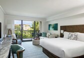A hotel room with a large bed, a desk with a chair, a balcony with a view of palm trees and buildings, and a painting on the wall.