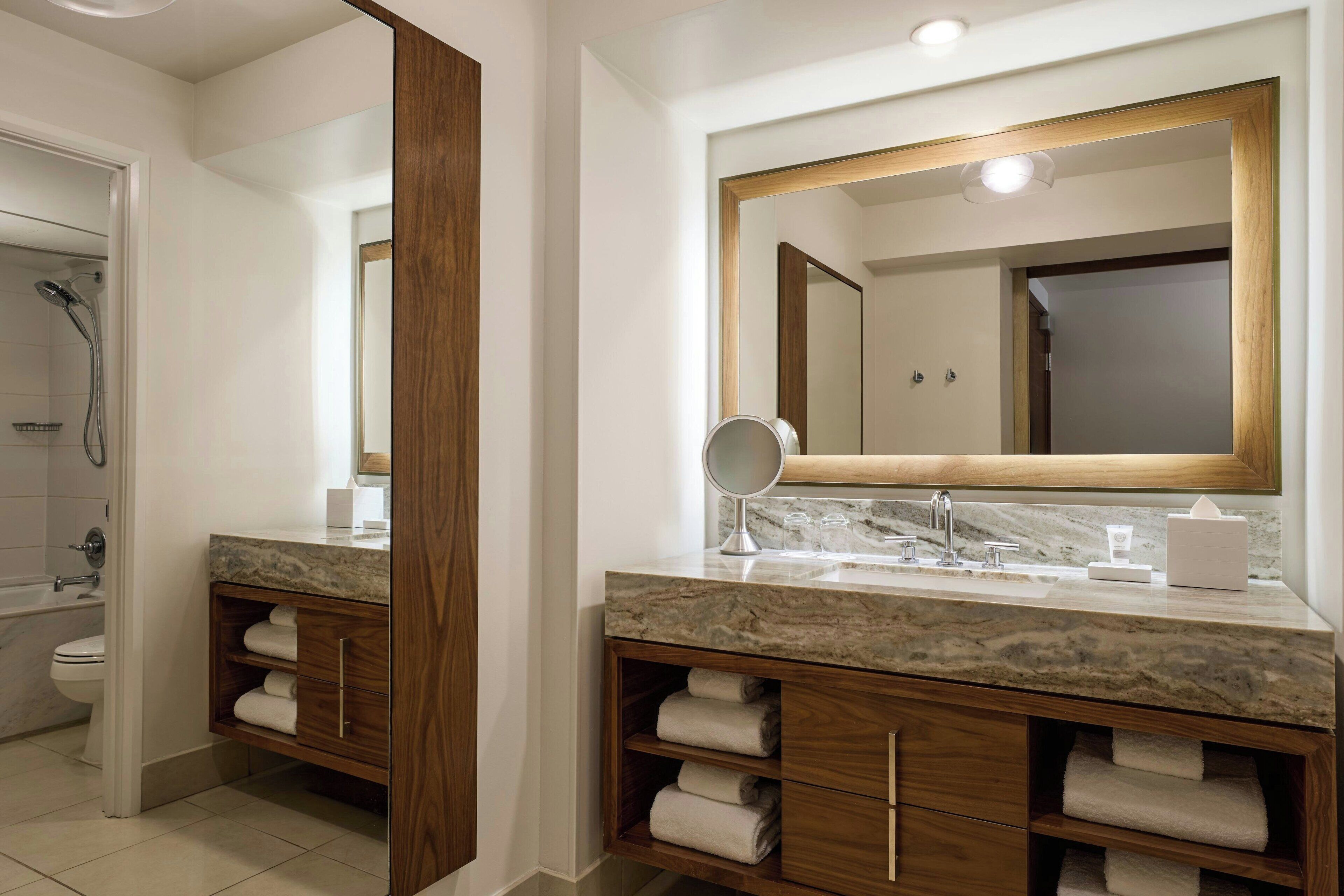 A modern bathroom with a marble vanity, wooden cabinetry, a large mirror, and a shower area.