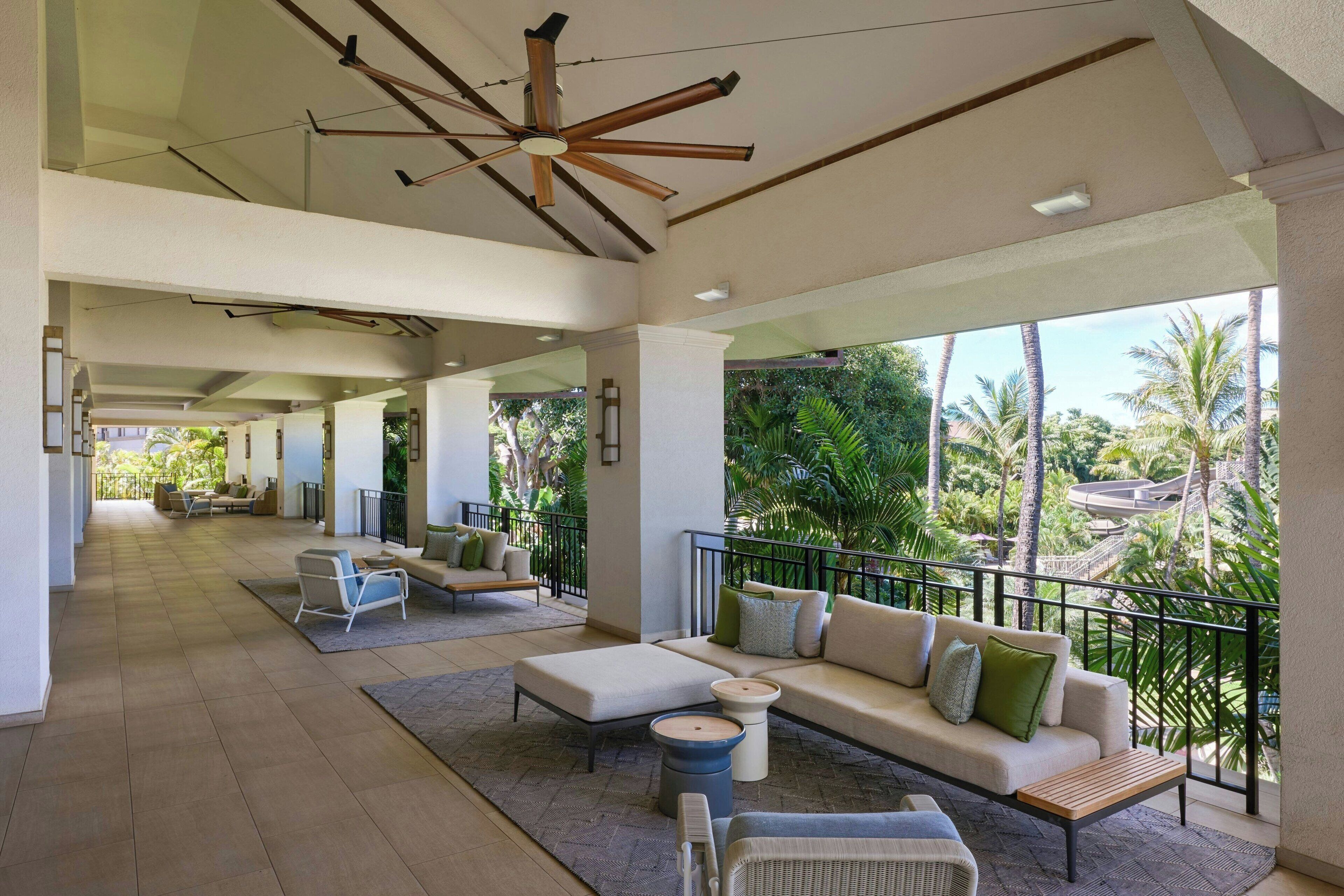 Un espace intérieur spacieux avec un balcon donnant sur des palmiers, comprenant un canapé, des fauteuils et un ventilateur de plafond.