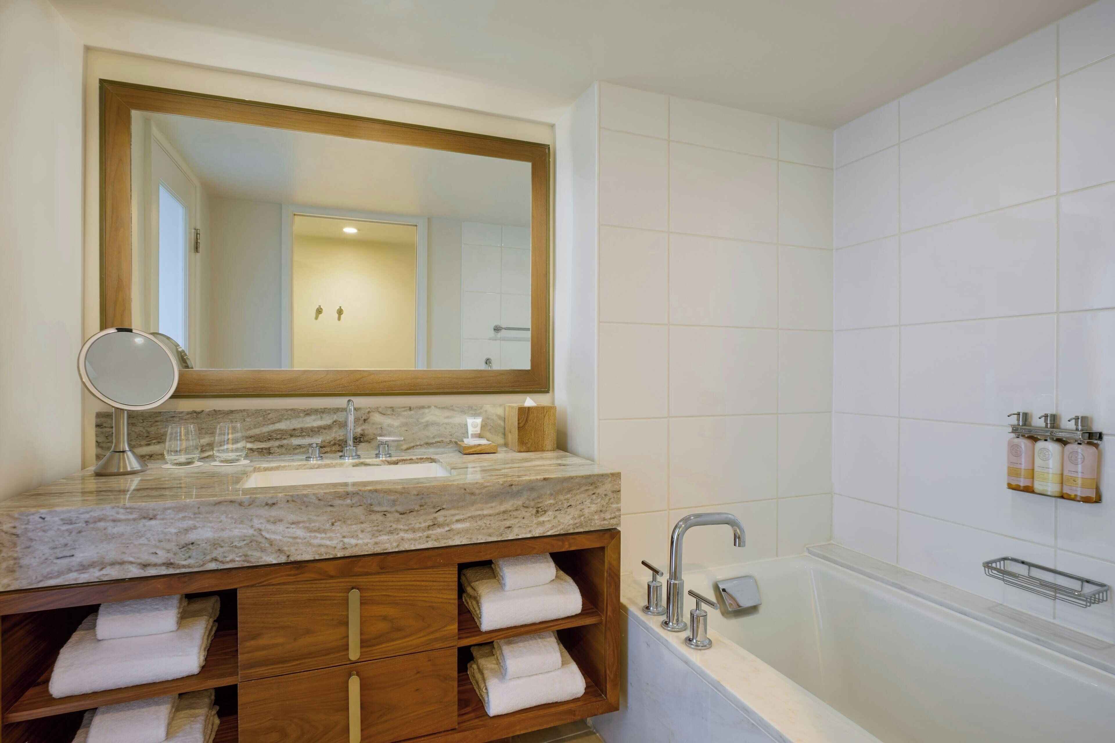 A bathroom with a marble sink, wooden vanity, a large mirror, a bathtub, and white tiled walls.