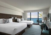 A hotel room with two beds, a desk, a chair, a TV, and a view of the ocean.