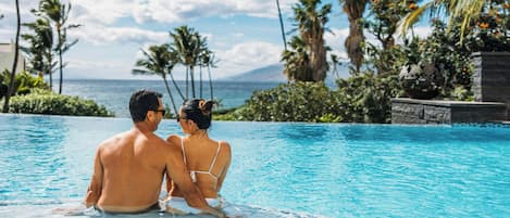 A couple sitting in a clear blue pool with a view of the ocean and palm trees.