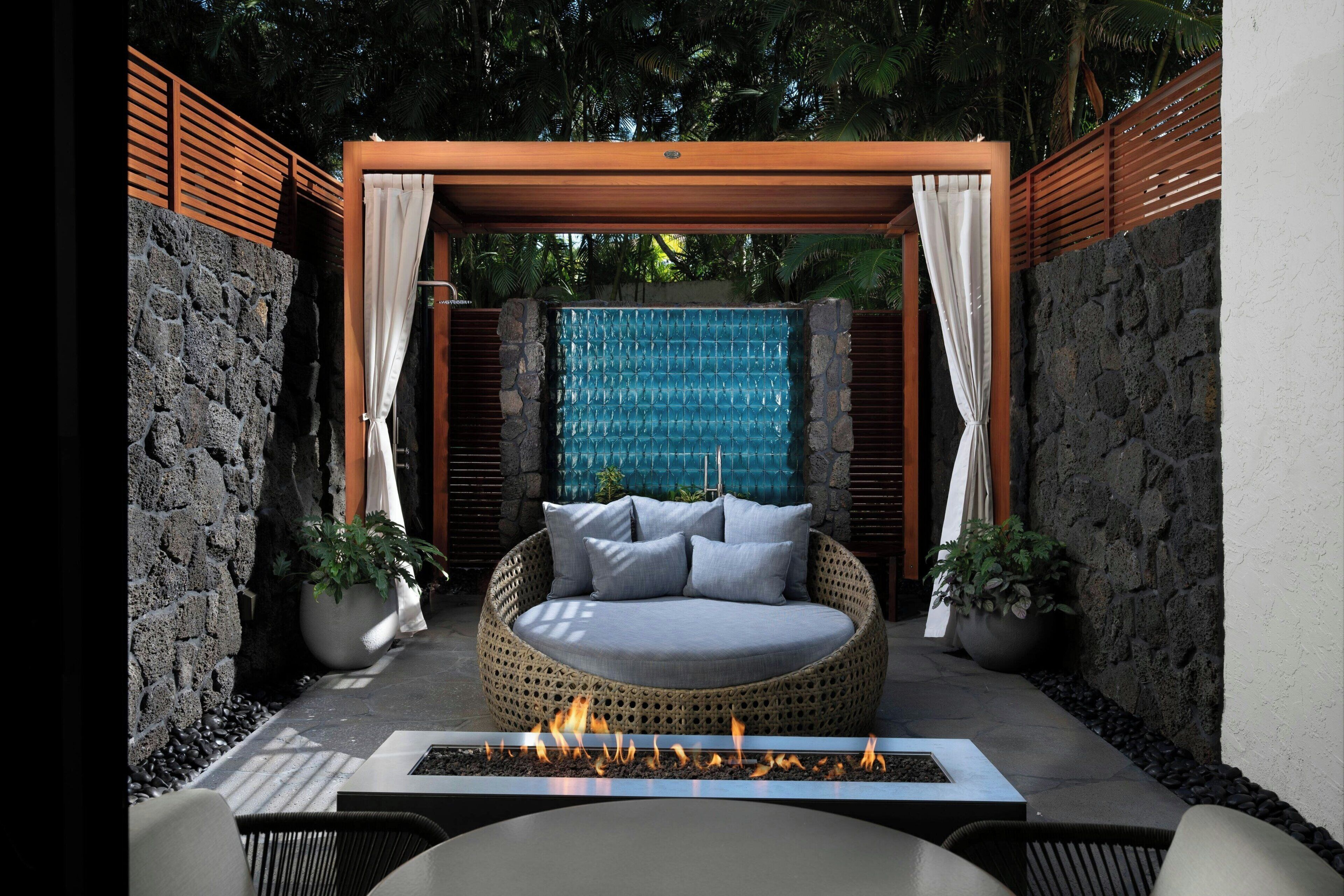 A round wicker sofa with cushions under a wooden pergola, a stone wall, and a small fire feature in the foreground.