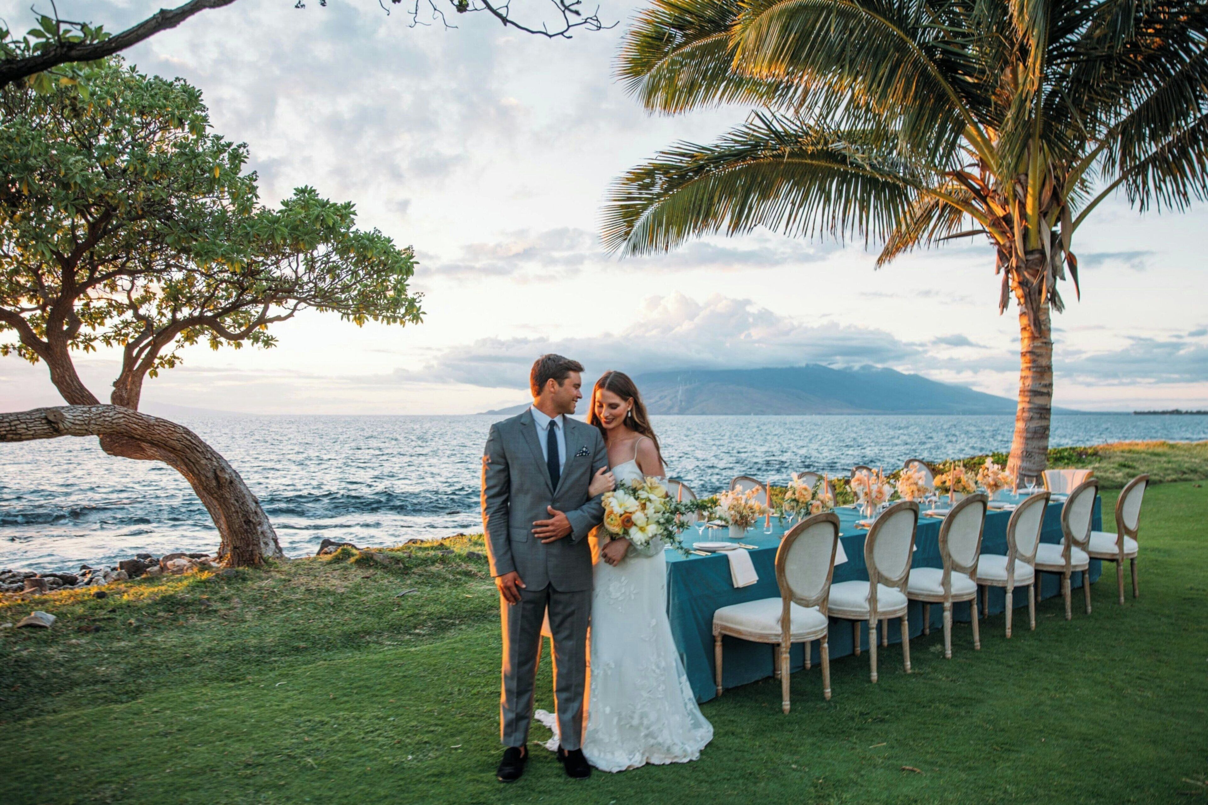 A couple standing by a table set for a wedding reception with a view of the ocean and mountains in the background.