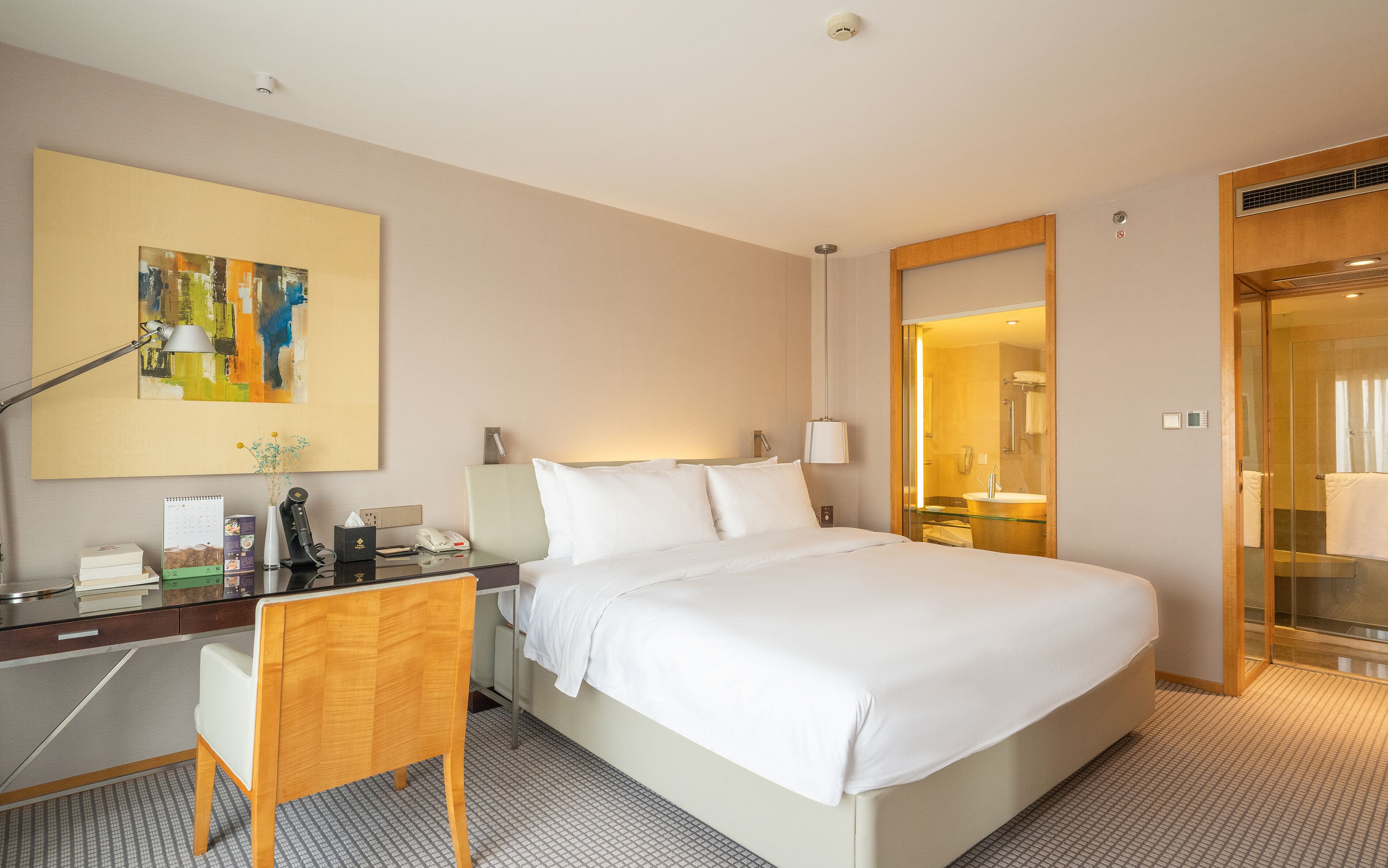 A hotel room with a bed, desk, chair, and a large painting on the wall.