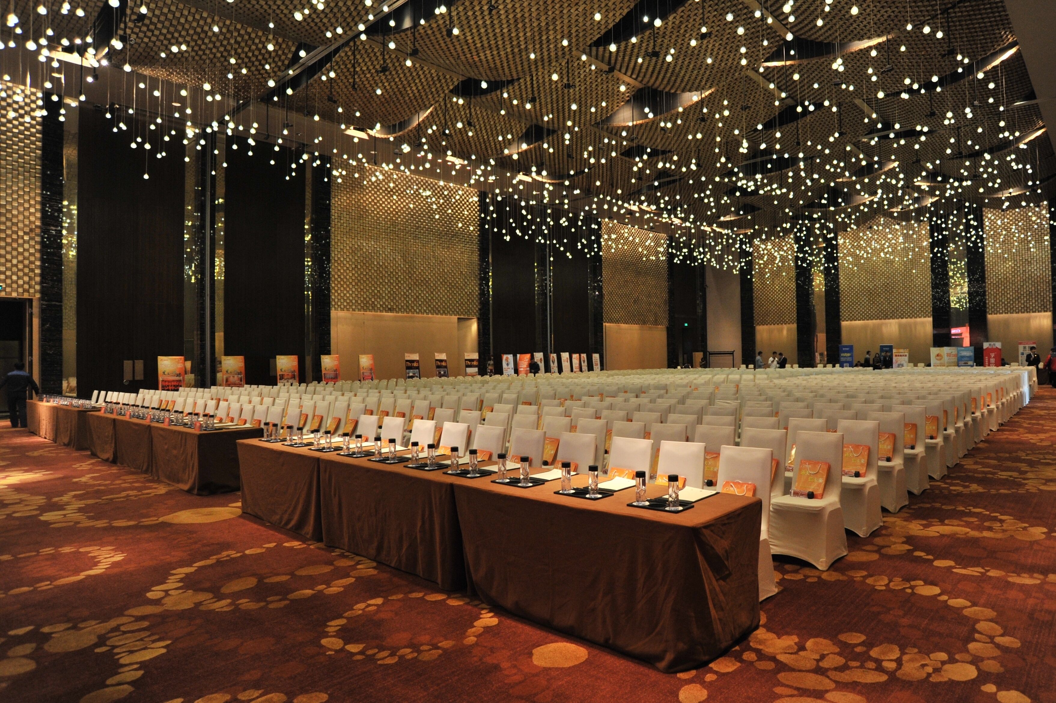 A large conference room with rows of white chairs, a long table with nameplates, and a ceiling adorned with numerous hanging lights.