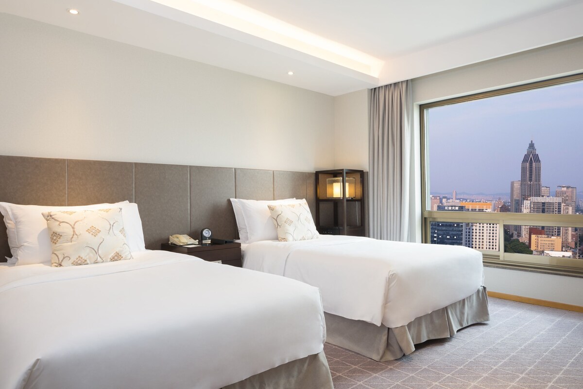 Habitación de hotel con dos camas, una mesita de noche, una lámpara y un amplio ventanal con vista a la ciudad.