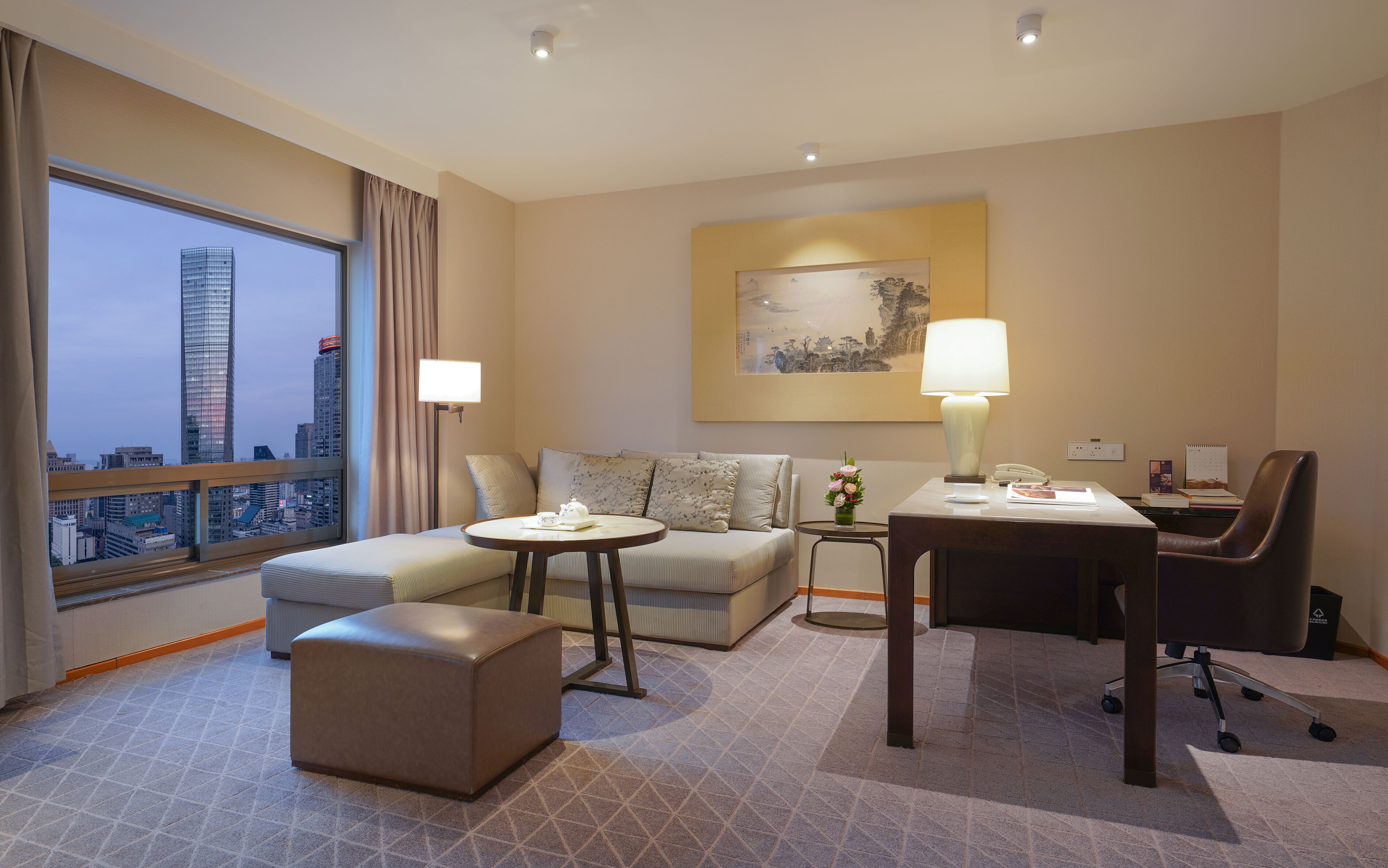 A hotel room with a sofa, ottoman, small table, and a desk with a chair. There is a window with a view of a cityscape and a painting on the wall.