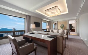 Une chambre d’hôtel dotée d’une grande fenêtre, d’un lustre, d’un ensemble de meubles de salon, d’un bureau et d’un téléviseur à écran plat.