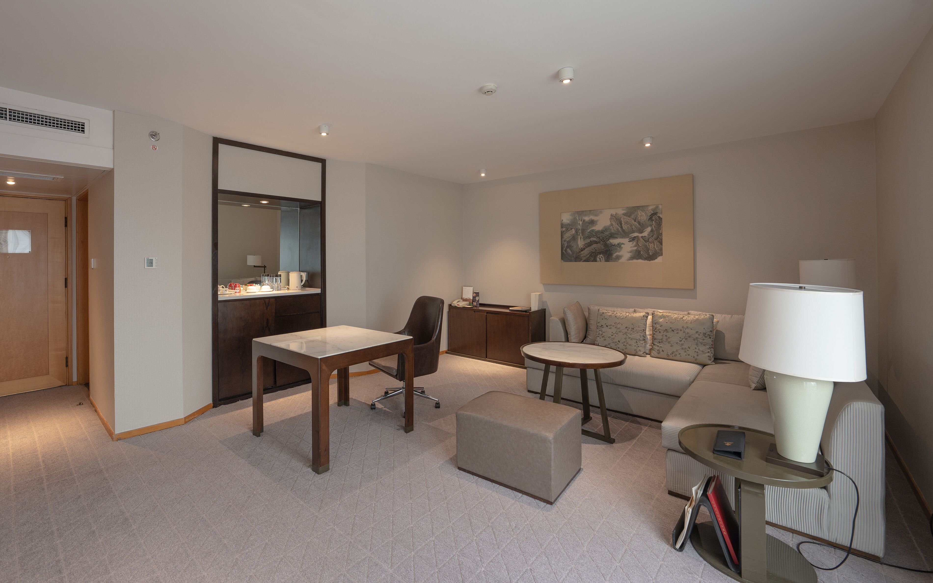 A modern hotel room with a wooden door, a desk with a chair, a sofa, a small round table, and a lamp.