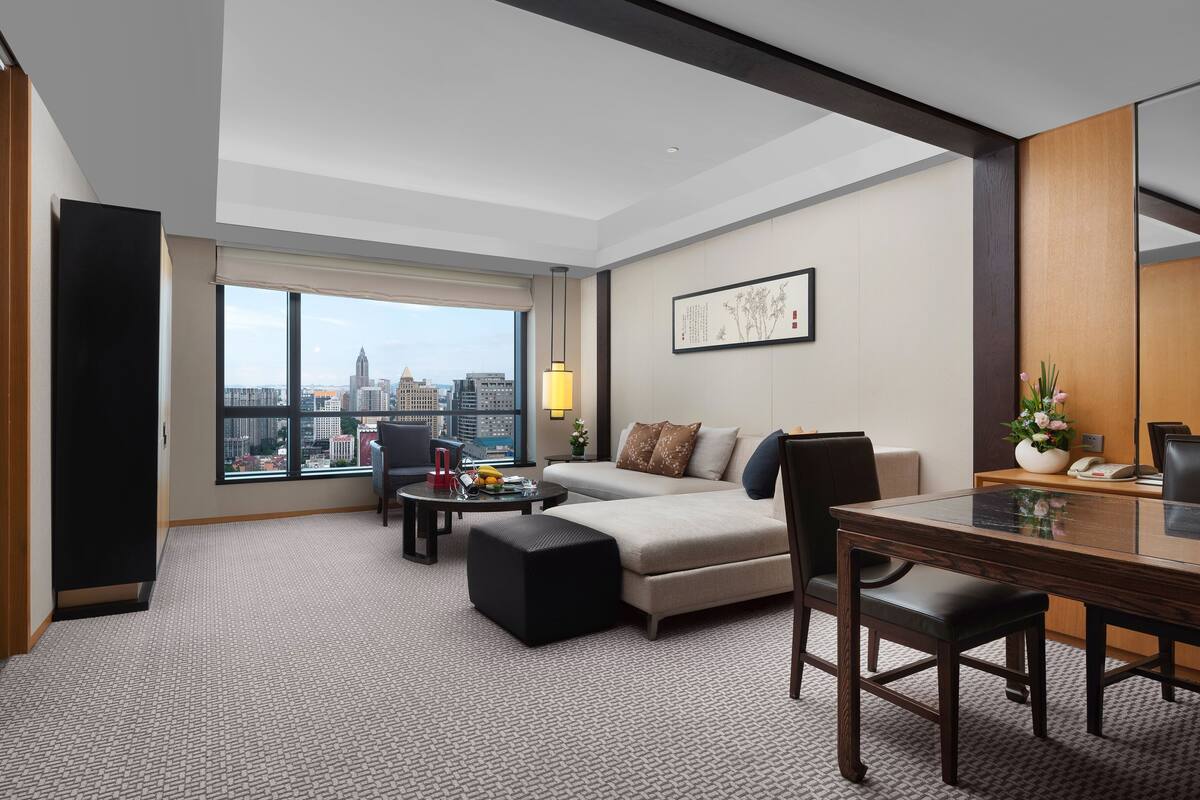 Une chambre d’hôtel moderne dotée d’une grande fenêtre donnant sur la ville, d’un canapé, d’une table à manger et d’une petite table d’appoint avec une lampe.