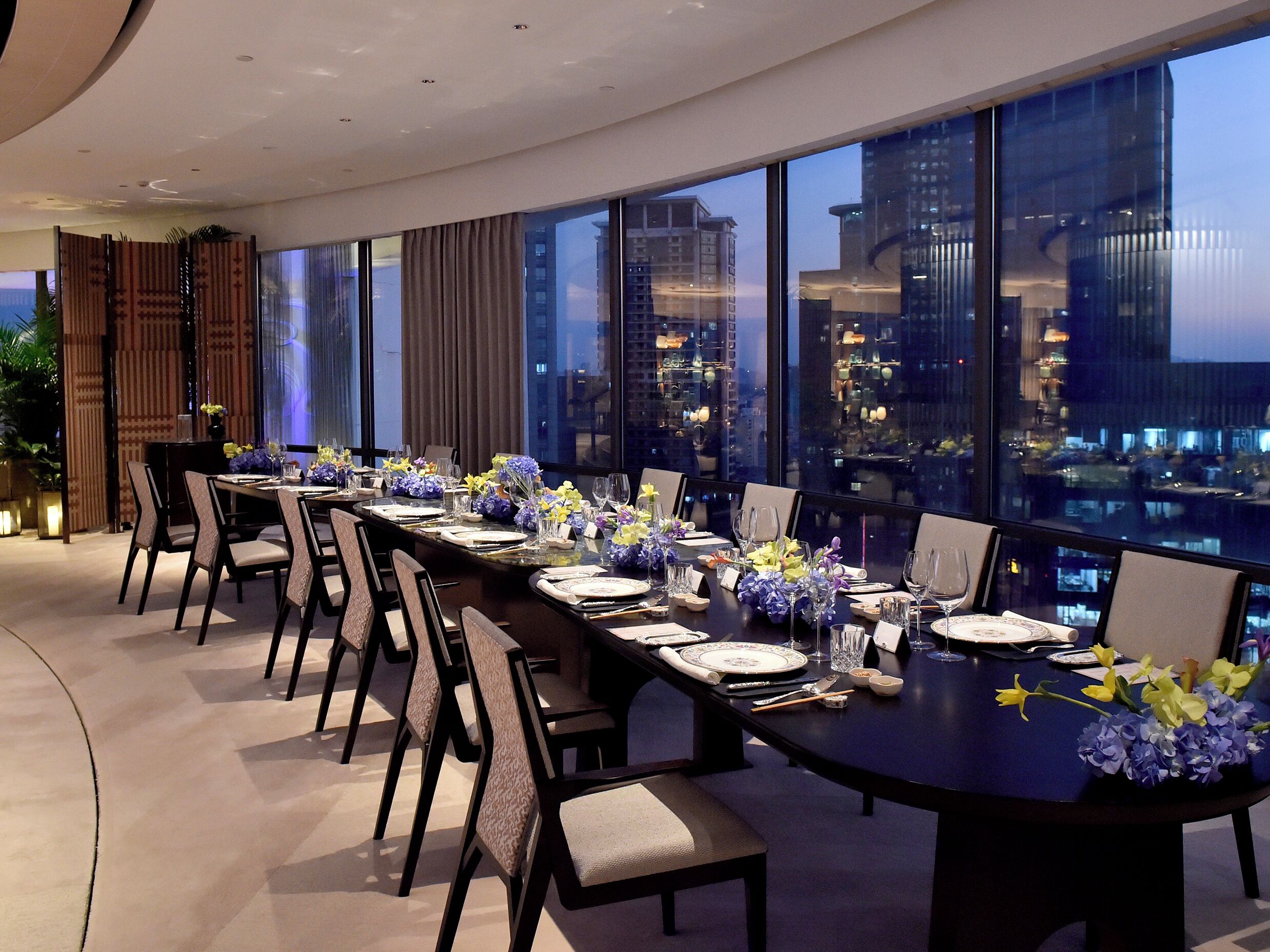Un comedor con una mesa larga dispuesta para una comida, ventanas amplias con vistas a la ciudad y mobiliario moderno.