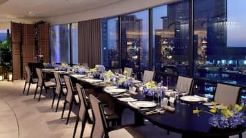 Un comedor con una mesa larga dispuesta para una comida, ventanas amplias con vistas a la ciudad y mobiliario moderno.