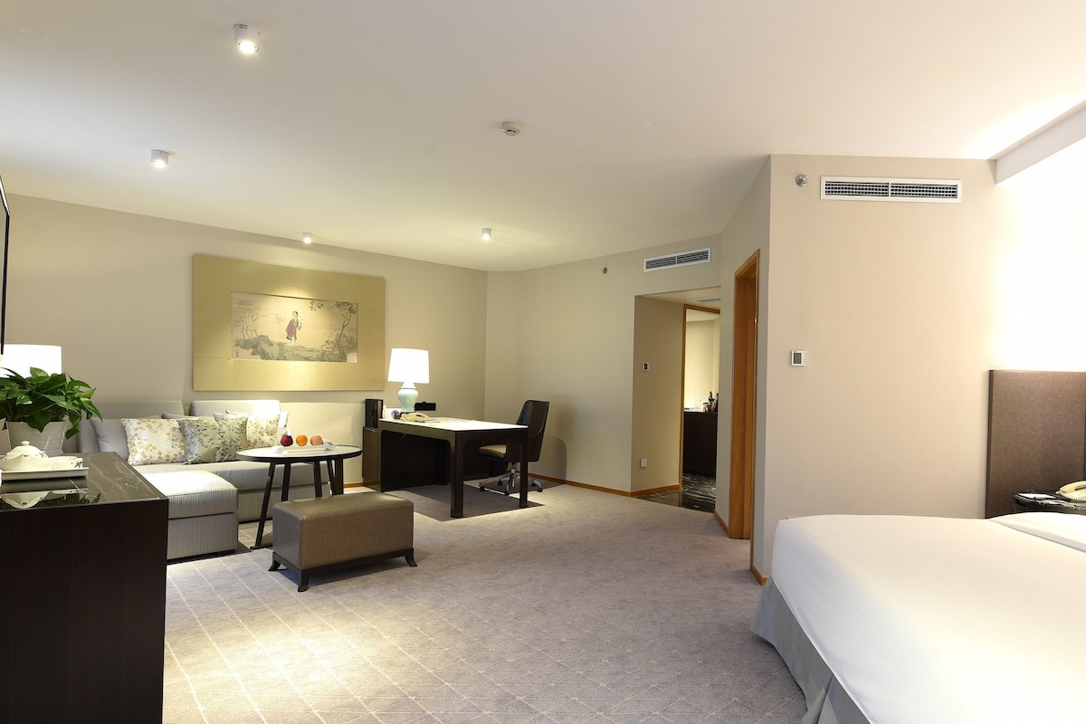 Una habitación de hotel con cama, escritorio, sillón, sofá, lámpara, planta y un cuadro en la pared.