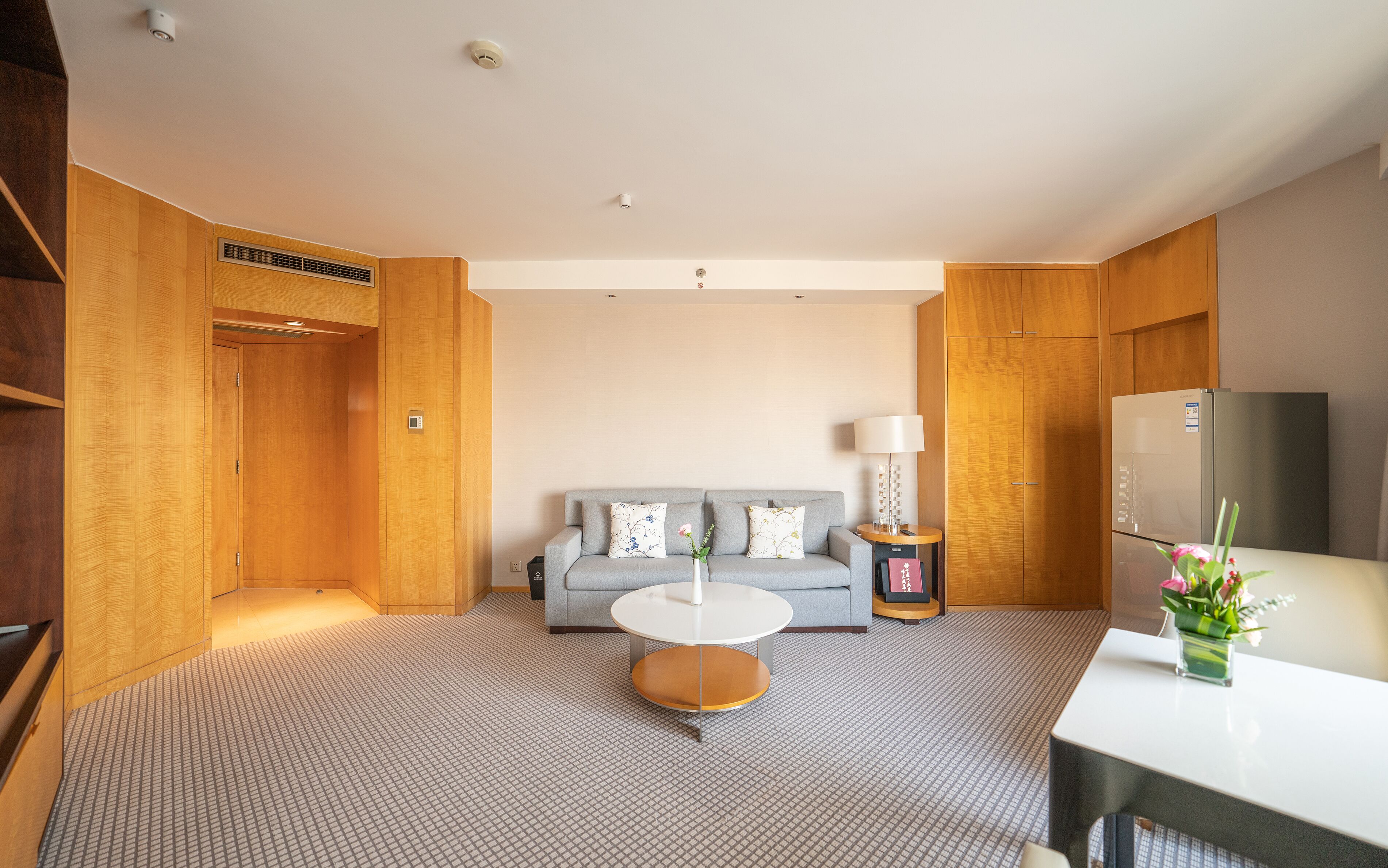 A hotel room with a grey sofa, a small round table, a white lamp, and a refrigerator.