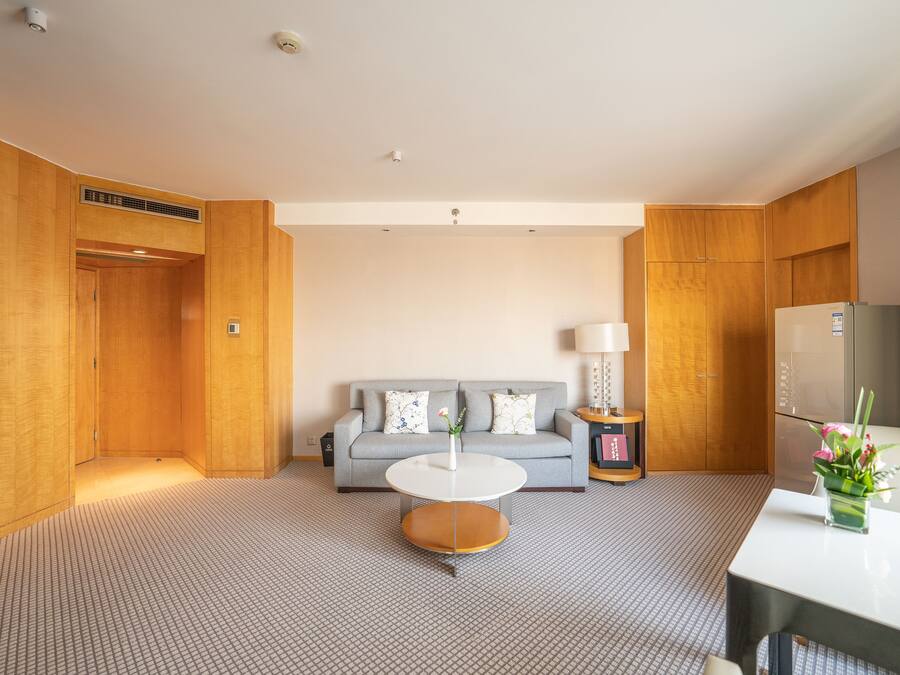 A hotel room with a grey sofa, a small round table, a white lamp, and a refrigerator.