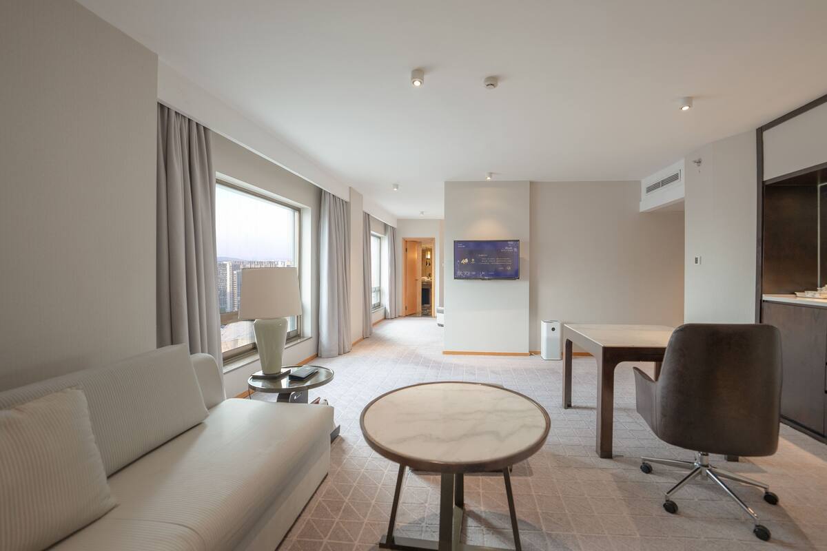 Une chambre d’hôtel équipée d’un canapé, d’une petite table ronde, d’une chaise, d’un téléviseur à écran plat fixé au mur et offrant une vue sur la ville par la fenêtre.