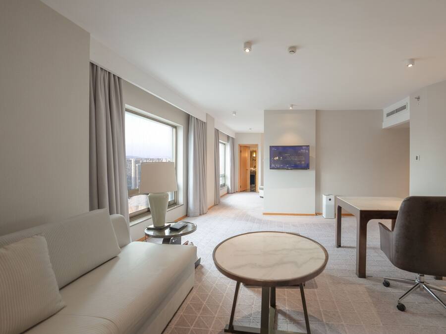 Ein Hotelzimmer mit einer Couch, einem kleinen runden Tisch, einem Sessel, einem an der Wand befestigten Flachbildfernseher und Blick auf die Stadt durch das Fenster.