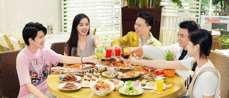 A group of people seated around a round table with various dishes and drinks, in an outdoor setting with plants and a white fence in the background.