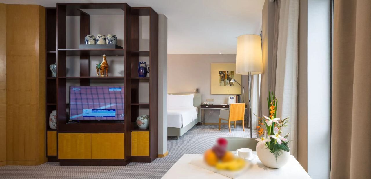 Habitación de hotel con televisor, escritorio, silla, cama, estantería y un jarrón con flores.