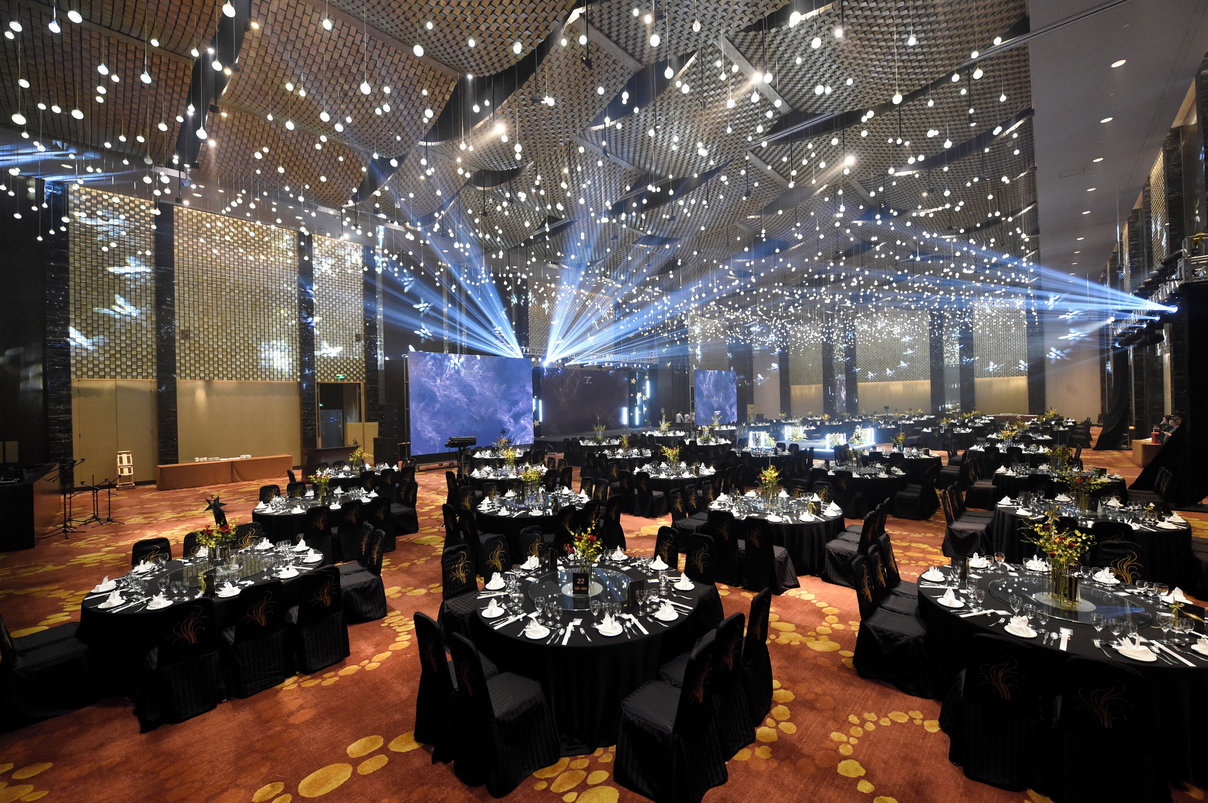 Un salón de banquetes con mesas redondas dispuestas para comer, un techo alto con luces colgantes y un escenario con un fondo azul.