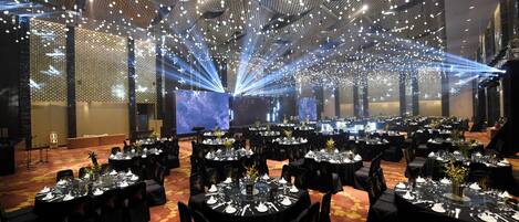 Un salón de banquetes con mesas redondas dispuestas para comer, un techo alto con luces colgantes y un escenario con un fondo azul.