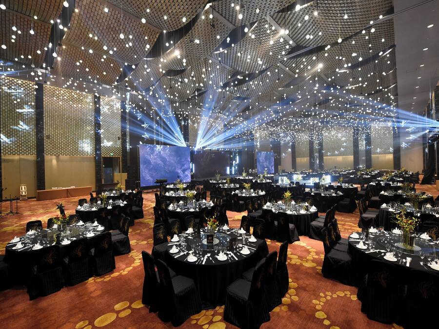 Una sala banchetti con tavoli rotondi apparecchiati per la cena, soffitto alto con luci a sospensione e un palco con sfondo blu.