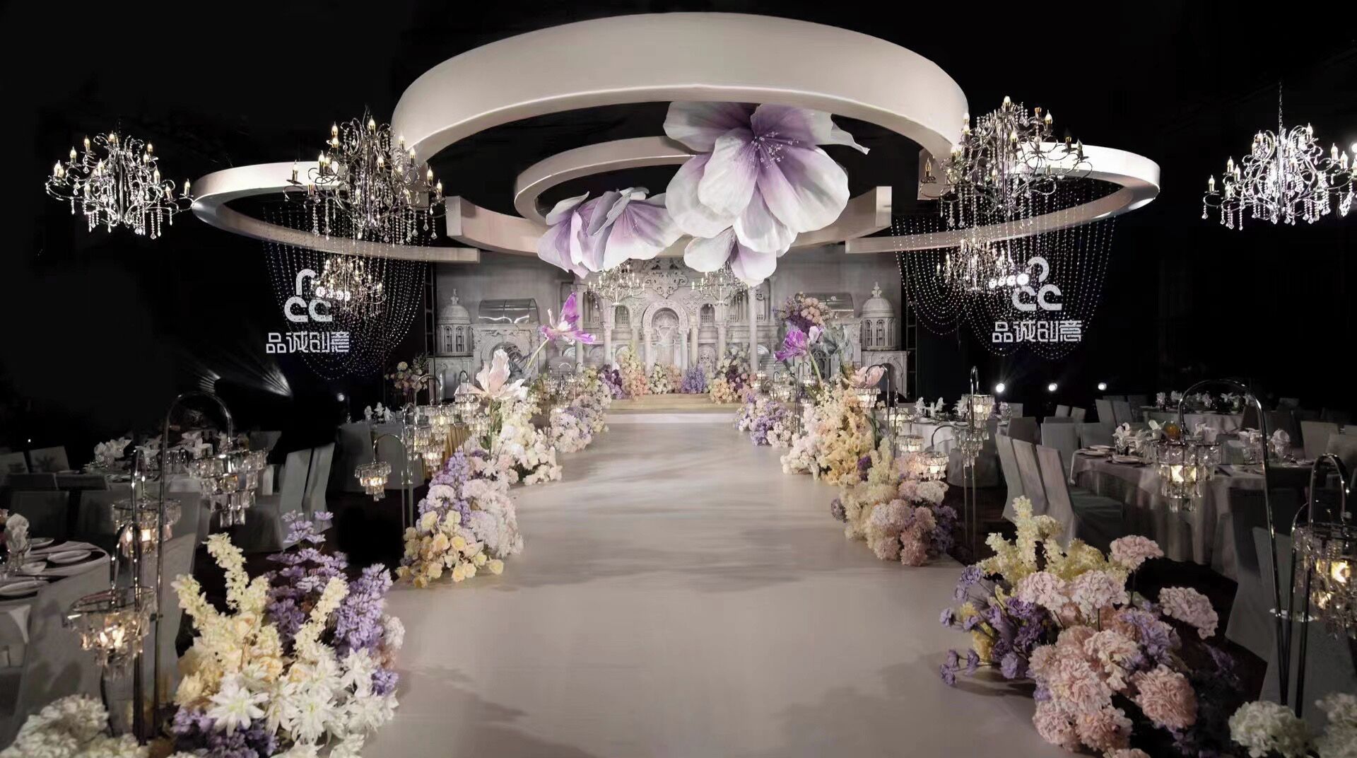 A formal event space with a central aisle, floral arrangements, and chandeliers.