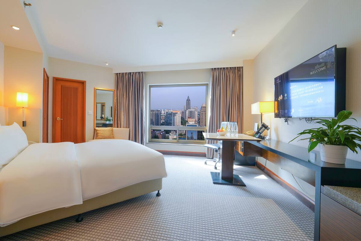 Une chambre d’hôtel avec un grand lit, un bureau doté d’un téléviseur à écran plat, une vue sur la ville et une plante en pot.