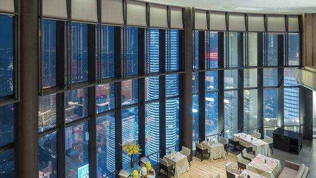 Habitación de hotel de gran altura con vistas panorámicas de la ciudad, varias mesas de comedor y una zona de estar moderna.