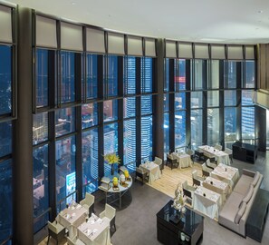 Habitación de hotel de gran altura con vistas panorámicas de la ciudad, varias mesas de comedor y una zona de estar moderna.
